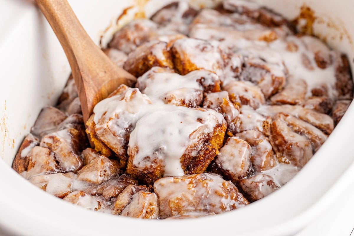 A wooden spoon scoops frosted cinnamon roll pieces from a white dish, with icing melted over the baked rolls.