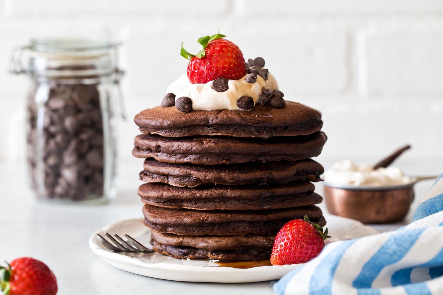 A stack of chocolate pancakes topped with whipped cream, chocolate chips, and a strawberry, with strawberries and a jar of chocolate chips in the background.