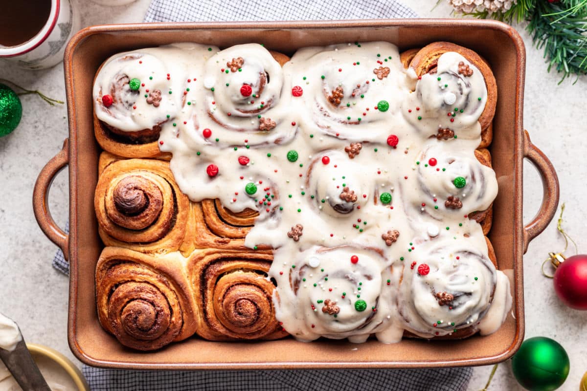 A baking dish filled with cinnamon rolls, half covered in white icing and decorated with red and green sprinkles, sits on a white surface with festive ornaments nearby.