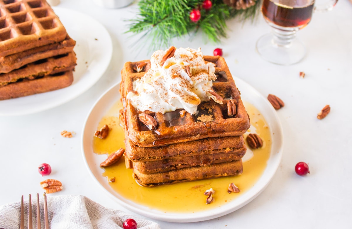 A stack of square waffles topped with whipped cream, pecans, and syrup sits on a white plate, surrounded by pecans and cranberries.