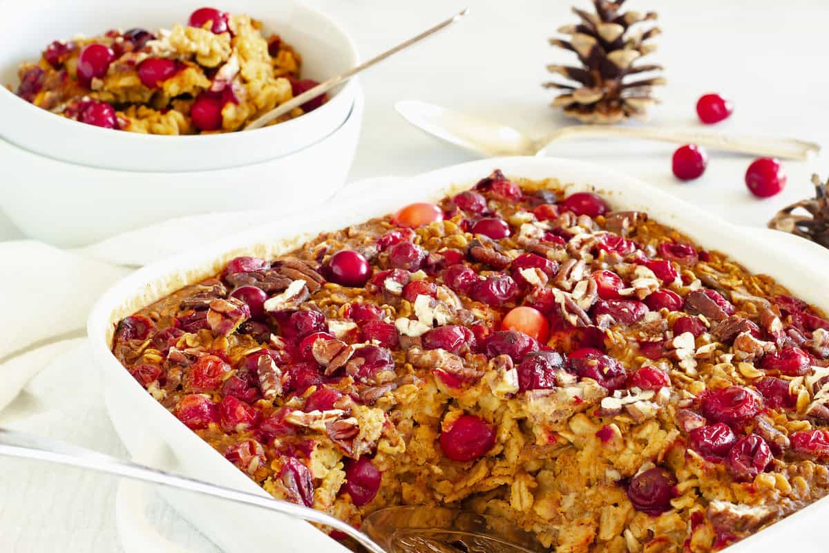 A baking dish of oatmeal topped with cranberries and pecans sits on a table, with a serving spoon and a bowl of the baked oatmeal nearby.