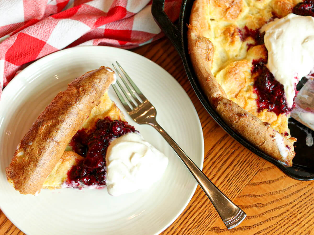 A slice of Dutch baby pancake with berry sauce and whipped cream on a plate, next to the remaining pancake in a skillet, with a fork and red-checkered cloth nearby.