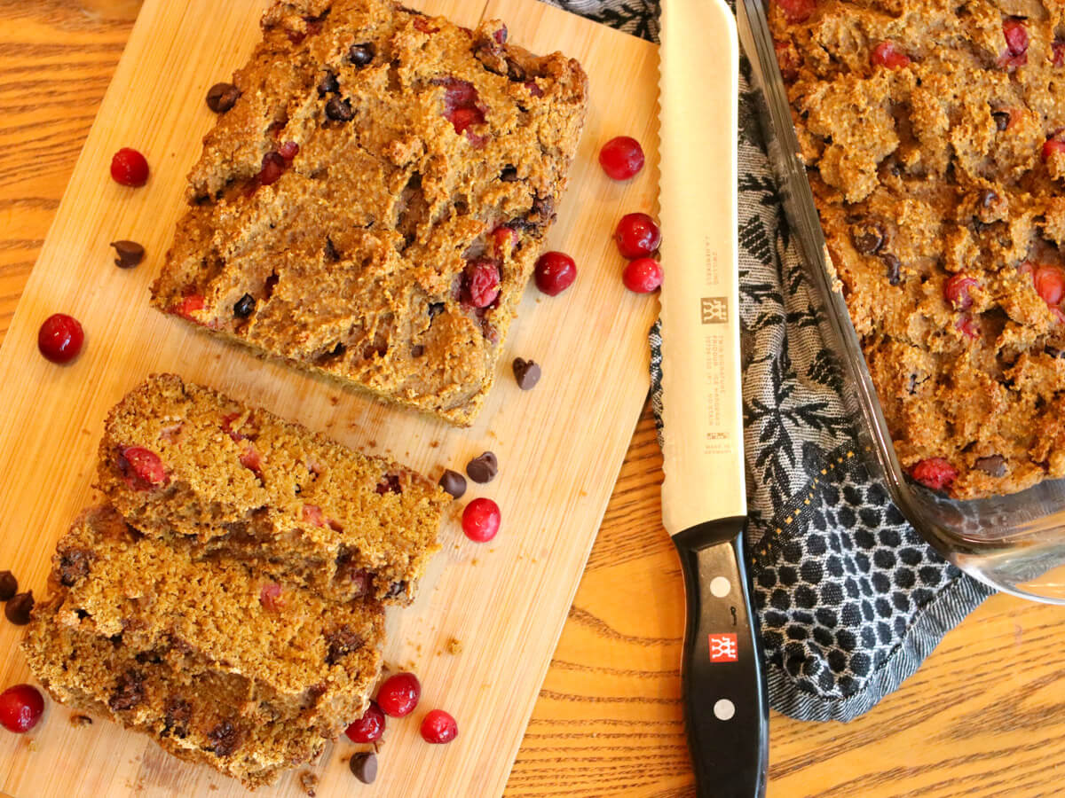 Sliced loaf of bread with cranberries and chocolate chips on a wooden cutting board, with a bread knife and a baking dish nearby.