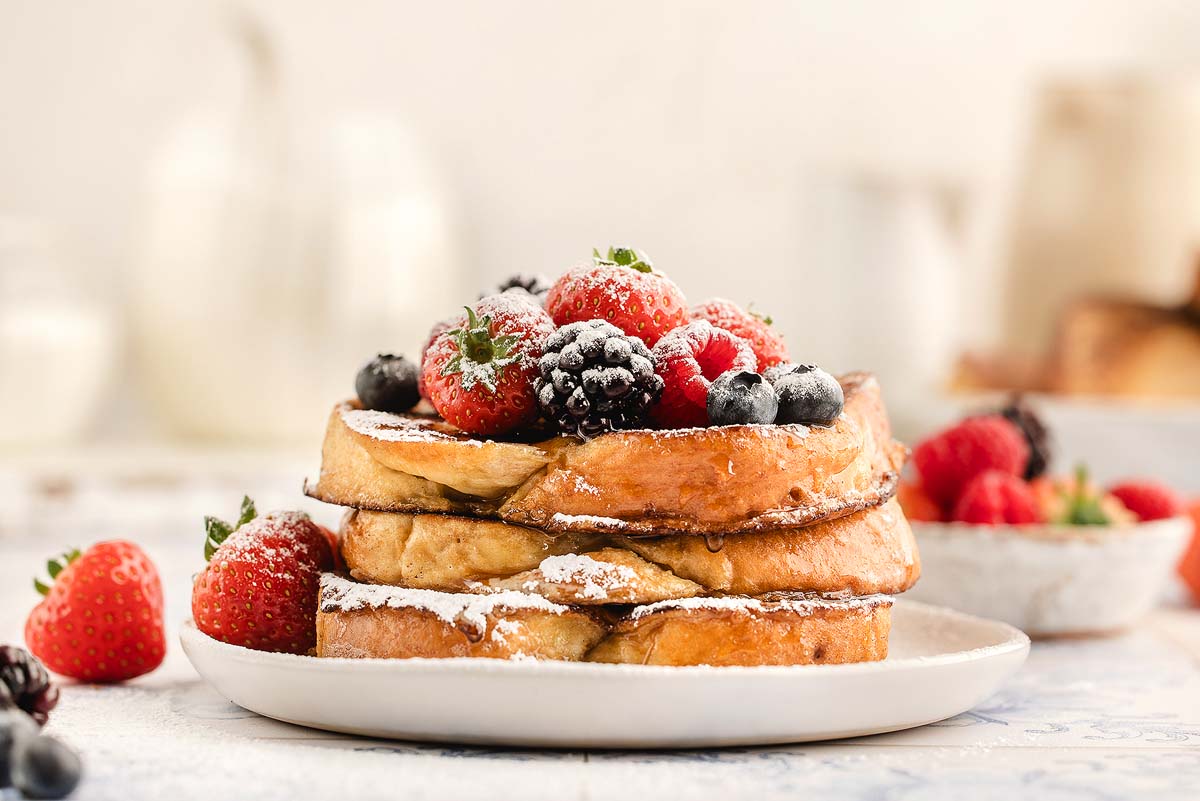 Three slices of French toast stacked on a white plate, topped with assorted berries and powdered sugar, with additional berries in the background.