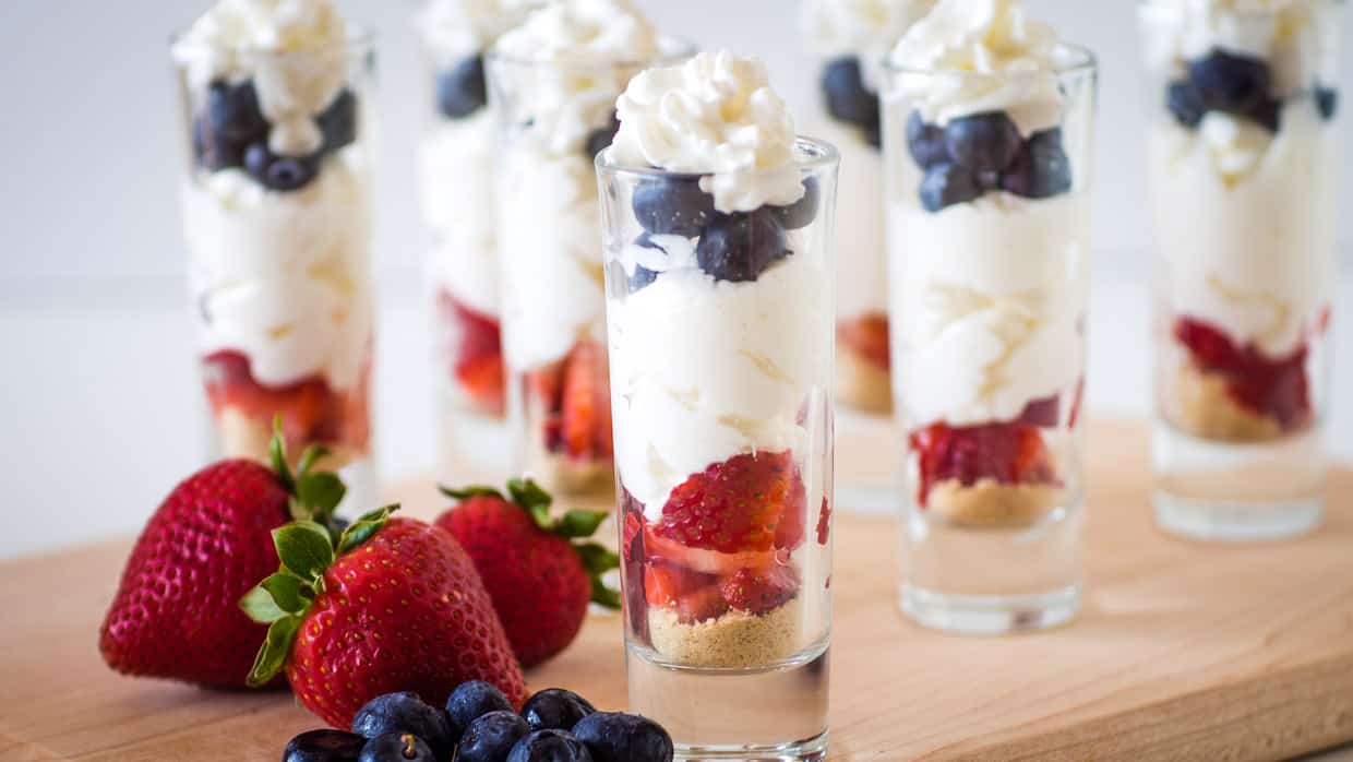 Several parfaits in shot glasses are arranged on a wooden board, layered with crumbs, strawberries, cream, and blueberries, with fresh berries placed in front.