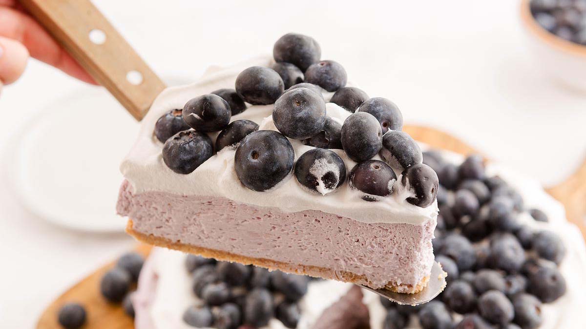 A slice of blueberry cheesecake with a graham cracker crust, topped with whipped cream and fresh blueberries, is being lifted from the whole cheesecake.