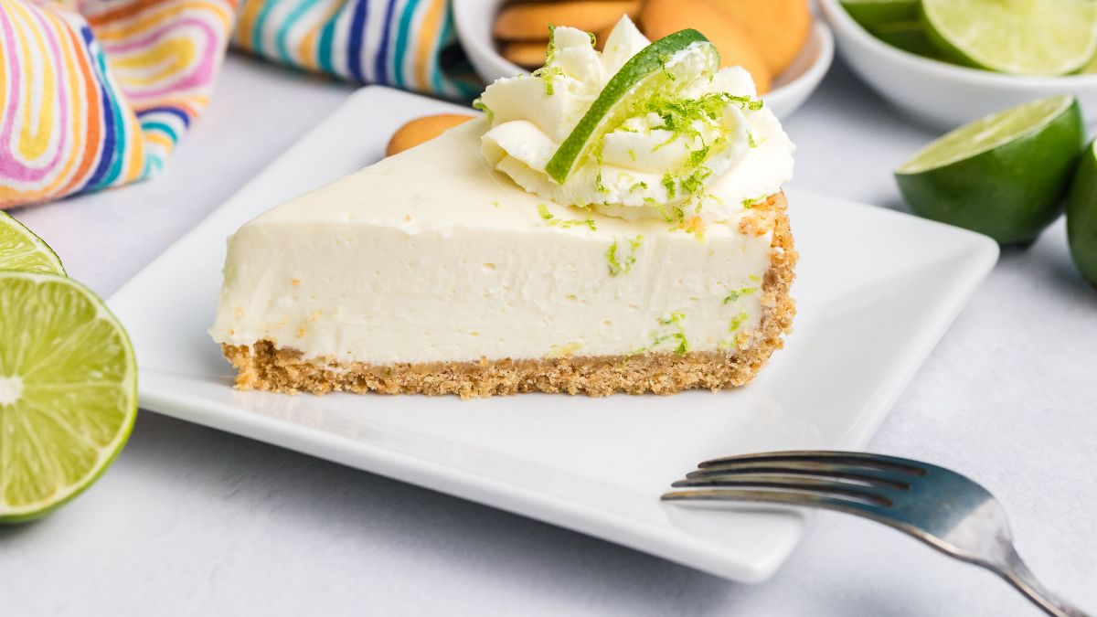A slice of key lime pie with graham cracker crust, whipped cream, and lime garnish on a white plate, surrounded by fresh limes and a fork.