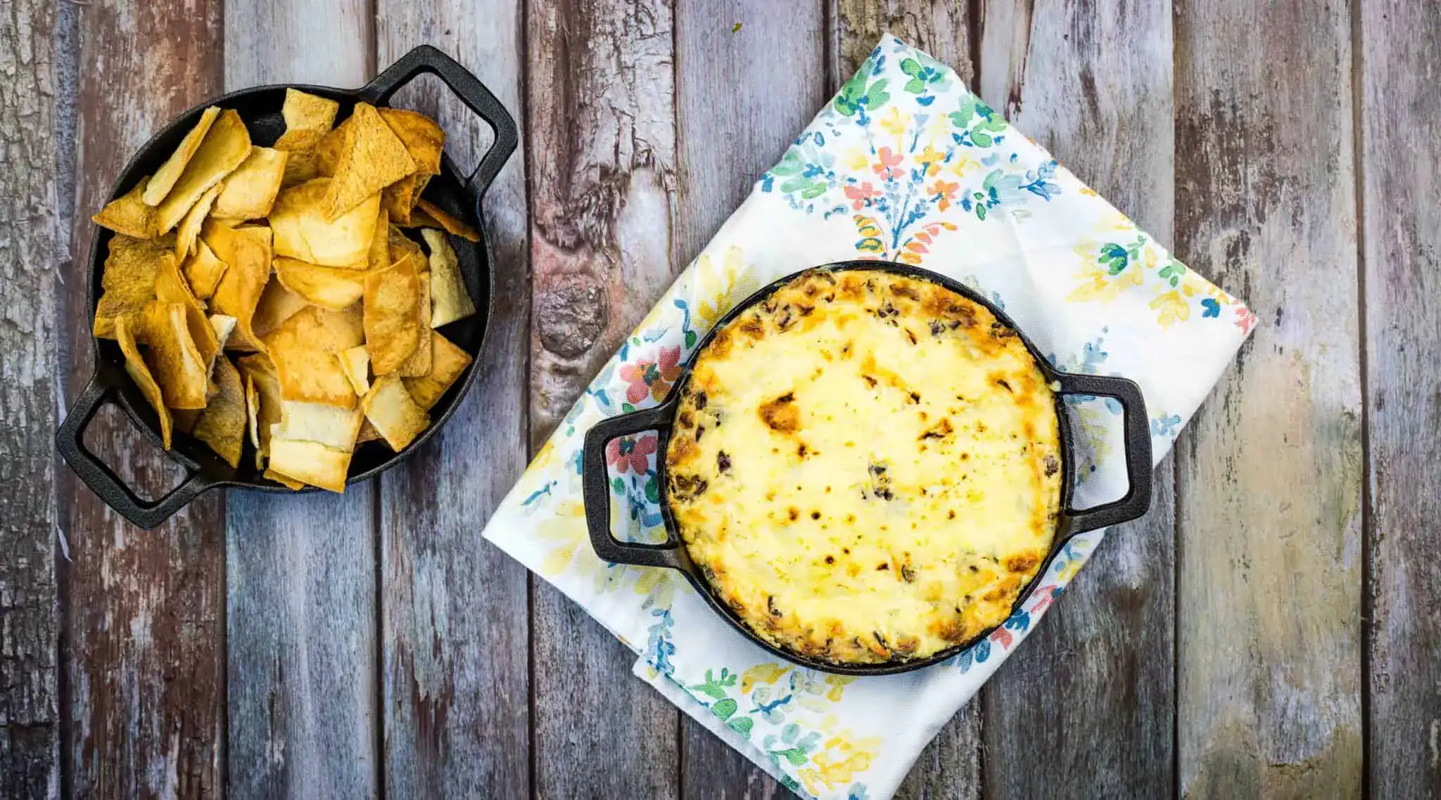 A small cast iron dish of baked cheese dip sits on a floral napkin next to a bowl of tortilla chips on a wooden surface.