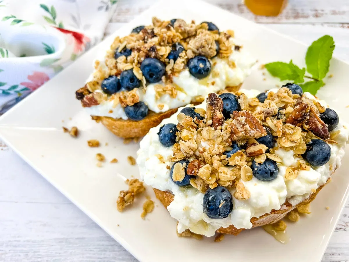 Two toasted bread halves topped with cottage cheese, blueberries, granola, and nuts are served on a white plate with a mint garnish.