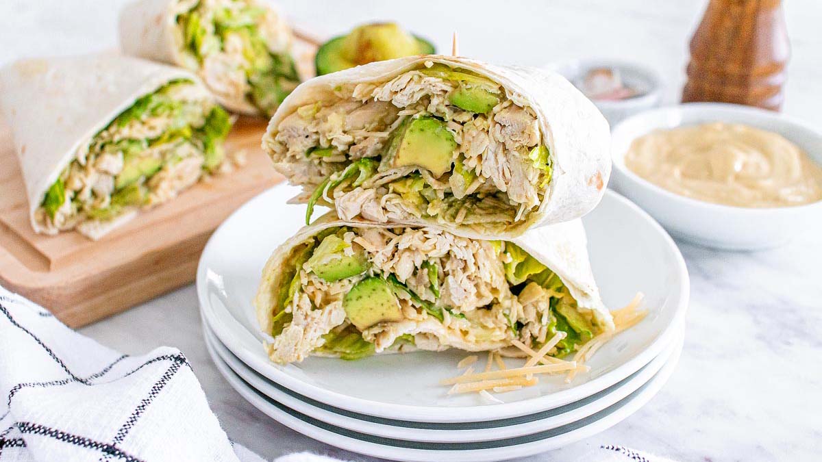 A chicken and avocado wrap sliced in half is stacked on a plate, with a bowl of sauce and a cutting board with more wraps in the background.