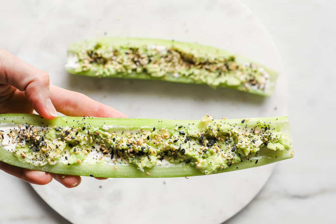 A hand holds a celery stick filled with cream cheese, avocado, and sprinkled with seasoning; another filled celery stick is on a plate in the background.