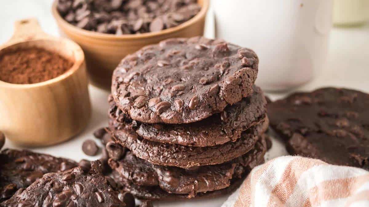 A stack of chocolate cookies with chocolate chips, surrounded by a bowl of chocolate chips, a wooden cup of cocoa powder, a chocolate cookie, and a glass of milk.