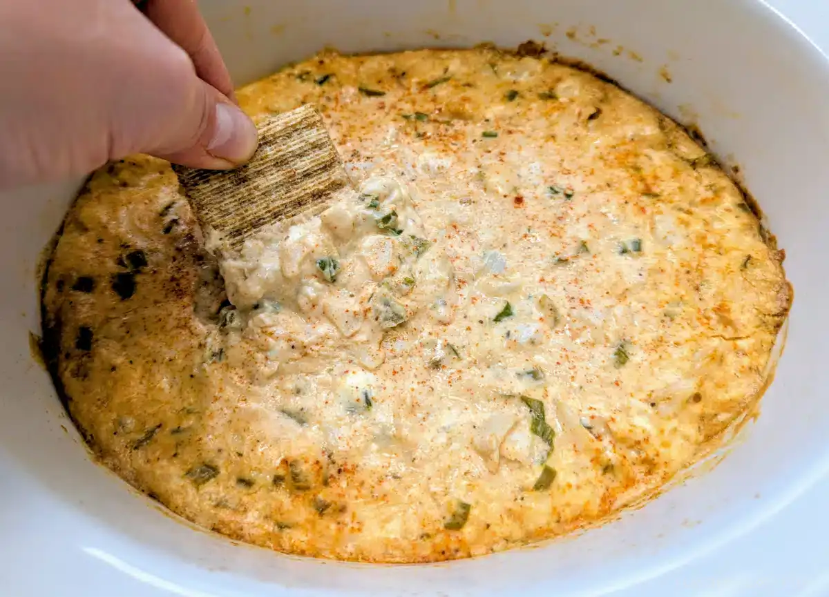 A hand dips a square cracker into a creamy, baked dip with visible herbs in a white dish.