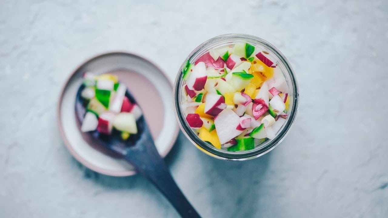 A jar filled with diced vegetables, including radishes, cucumbers, and yellow peppers, sits next to a spoon holding a similar mix on a small plate.
