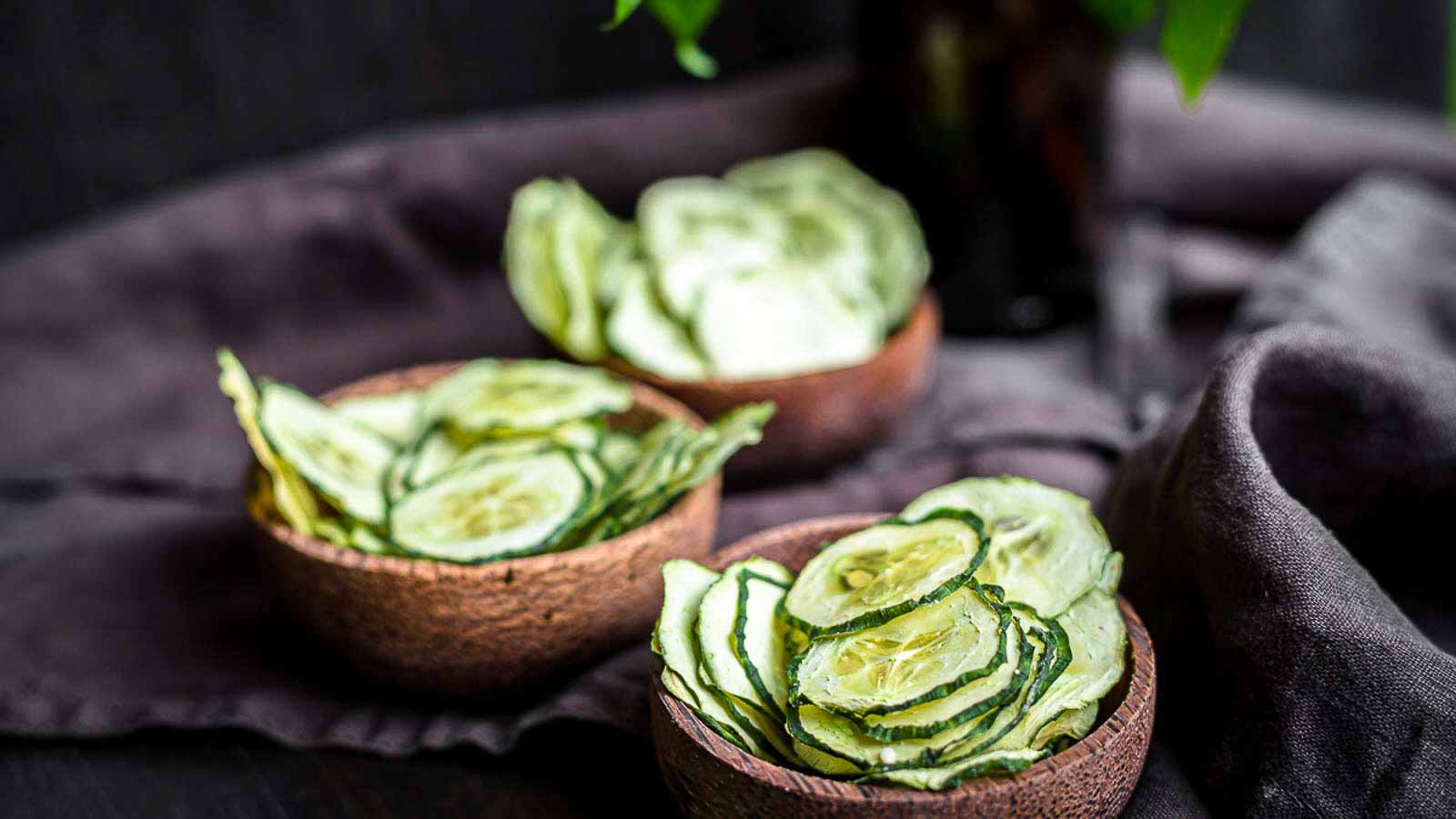Two small bowls filled with thinly sliced cucumber chips are placed on a dark cloth surface.