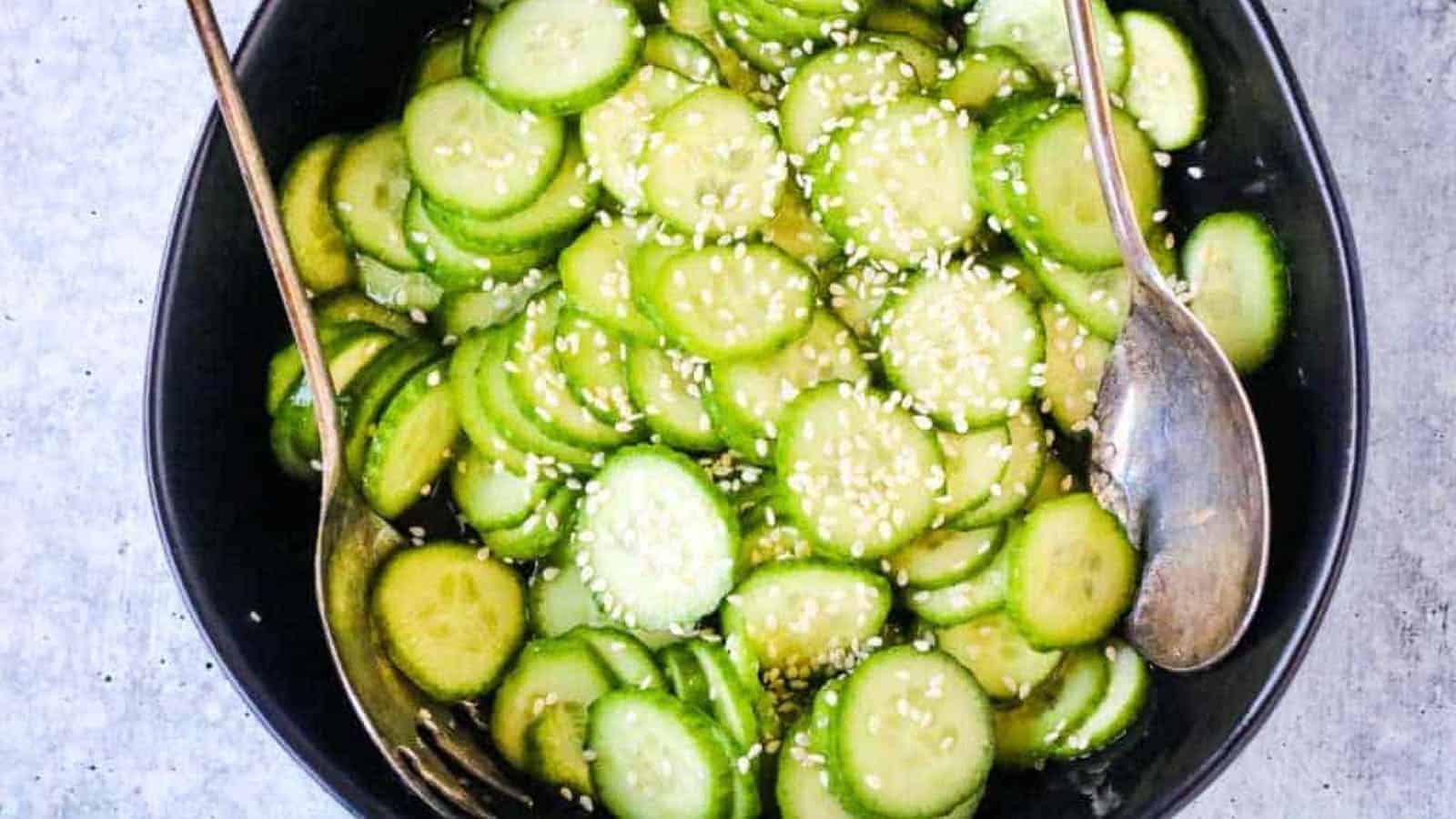 A black bowl filled with sliced cucumbers topped with sesame seeds, with a fork and spoon placed inside the bowl.