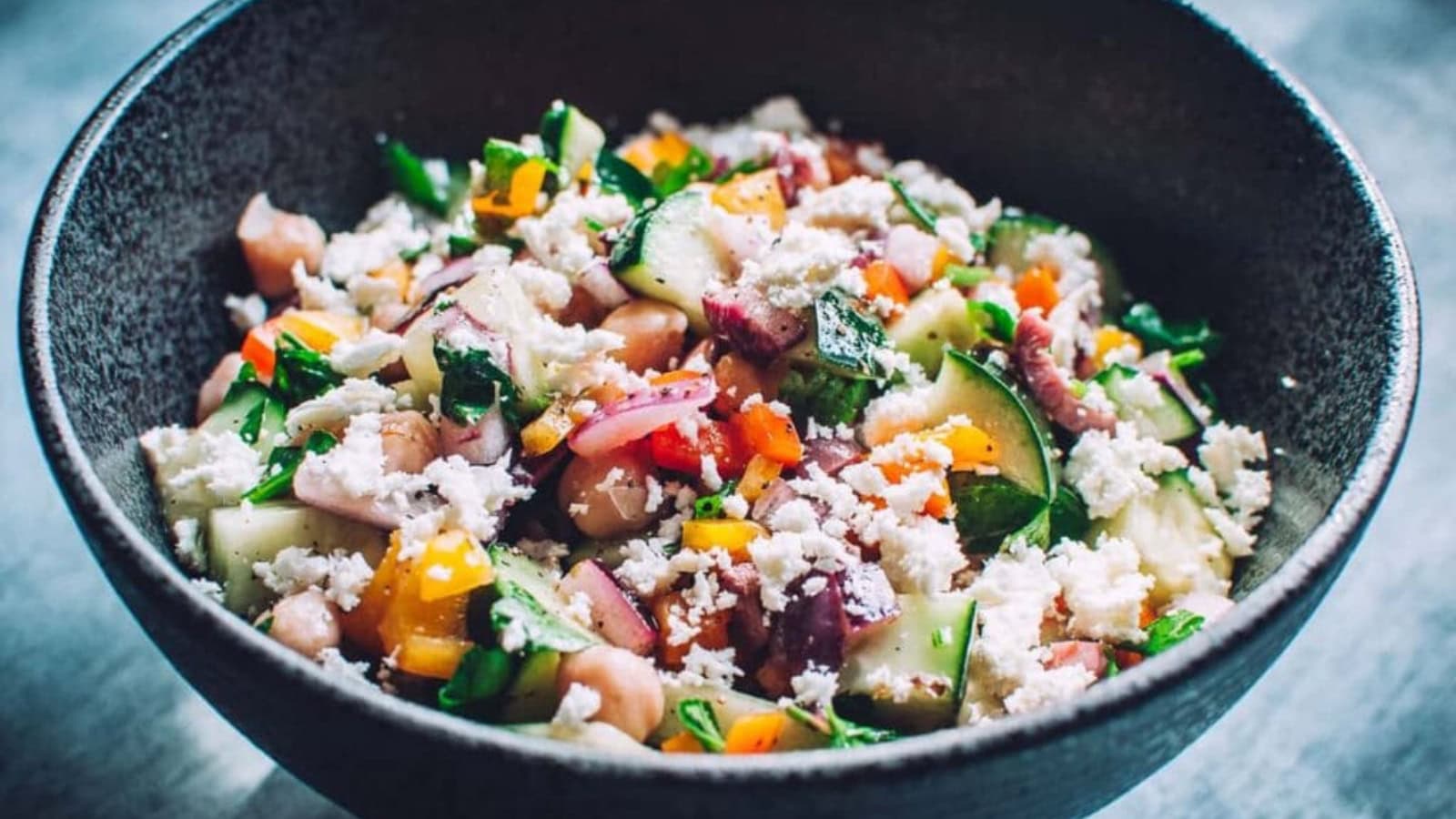 A black bowl filled with a mixed salad containing chopped cucumbers, tomatoes, beans, red onions, herbs, and crumbled cheese.