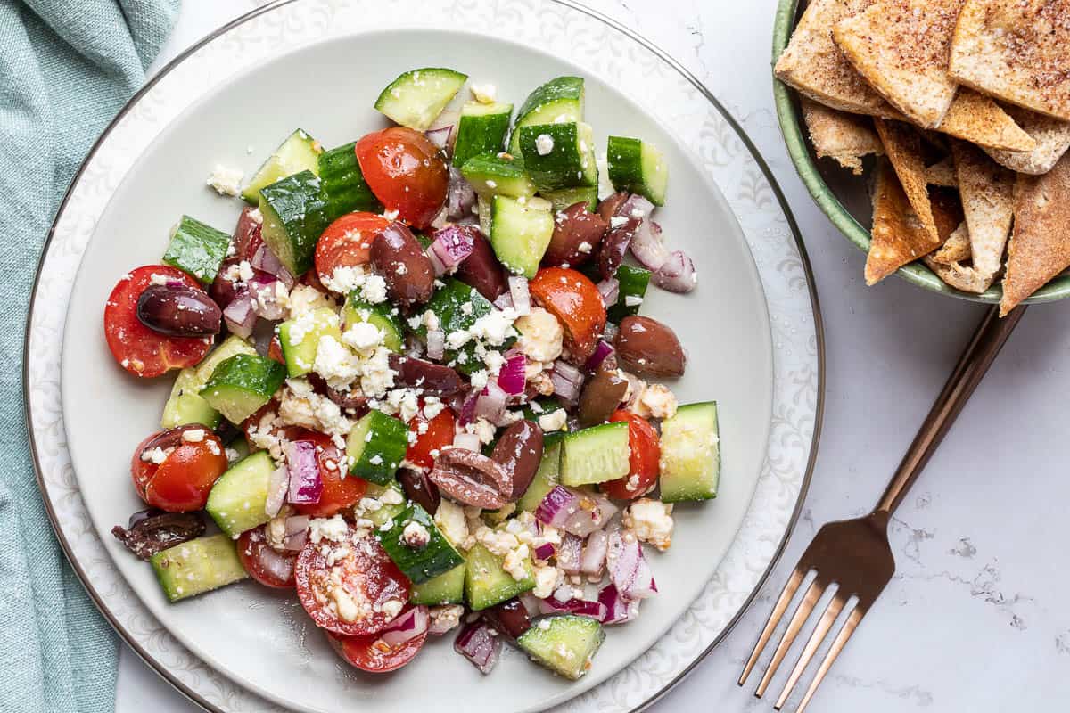 A plate of Greek salad with cucumbers, cherry tomatoes, red onions, Kalamata olives, and feta cheese next to a bowl of pita chips and a fork.