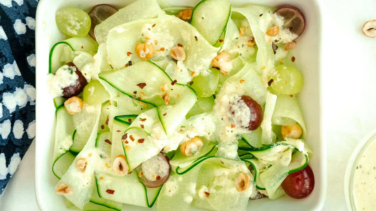 A salad with thinly sliced cucumber ribbons, red and green grapes, hazelnuts, cheese crumbles, and a sprinkle of red pepper flakes on a white plate.