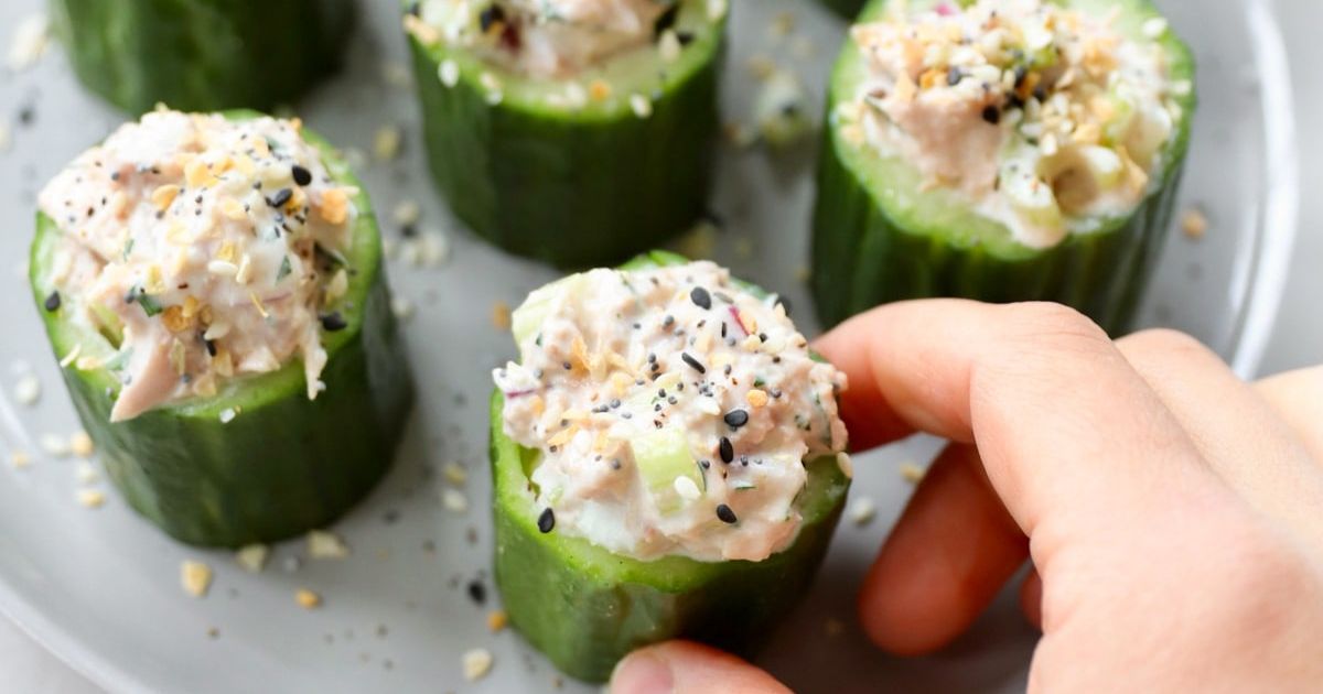 A hand reaches for a cucumber cup filled with tuna salad and sprinkled with seasoning, arranged on a white plate.