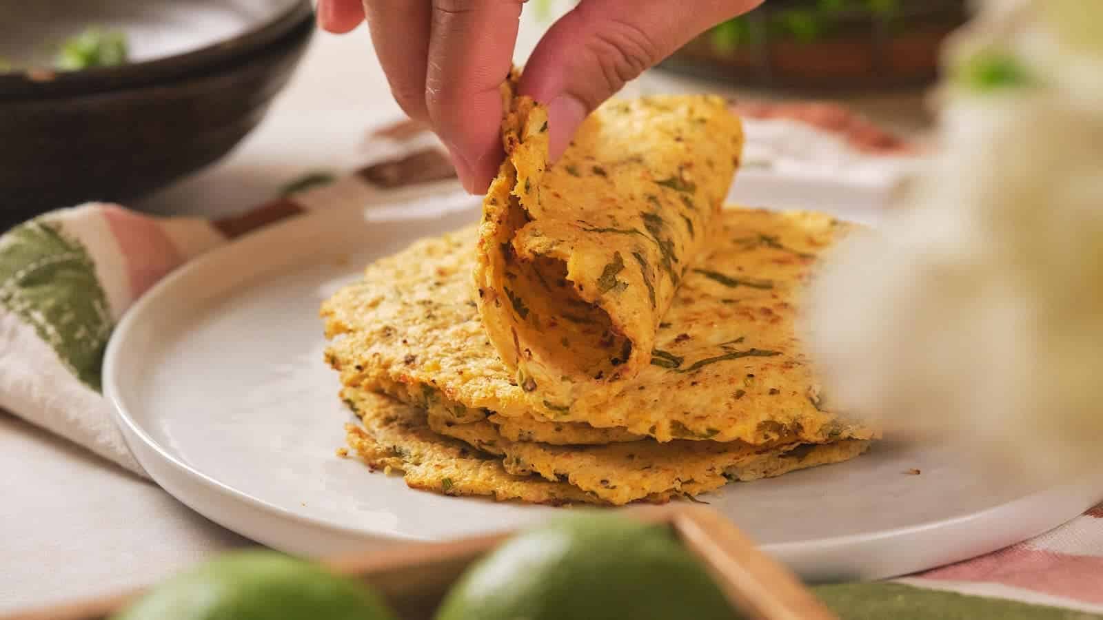 A hand lifts a folded flatbread with herbs from a stack on a white plate, surrounded by limes and a green-patterned napkin.