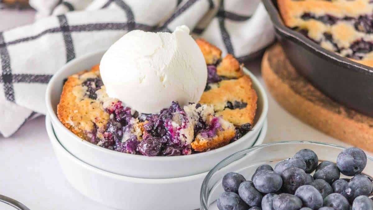 A bowl of blueberry cobbler topped with a scoop of vanilla ice cream, with fresh blueberries and a checkered cloth in the background.