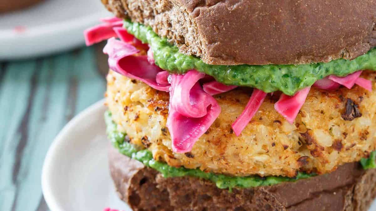 Close-up of a veggie burger with a brown bun, green sauce, pink pickled onions, and a golden vegetable patty on a white plate.