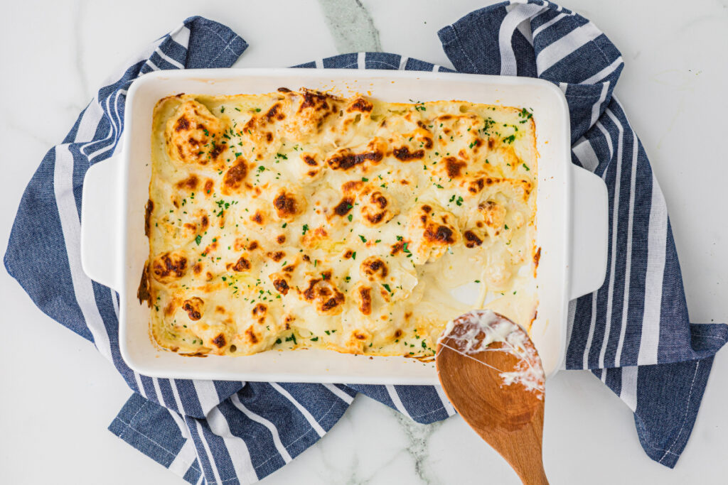 A baked casserole with a golden, bubbly cheese topping in a white dish, placed on a striped blue towel, with a wooden spoon beside it.