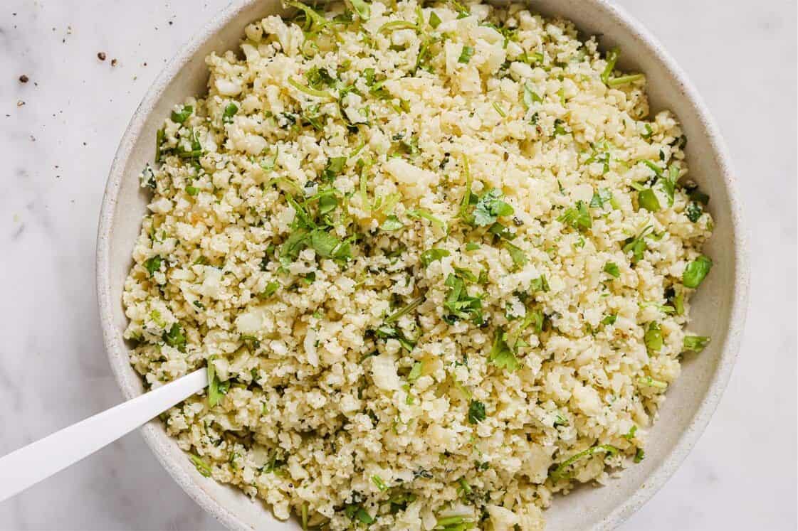 A bowl of cauliflower rice mixed with chopped herbs, with a white spoon resting inside the bowl, on a white surface.