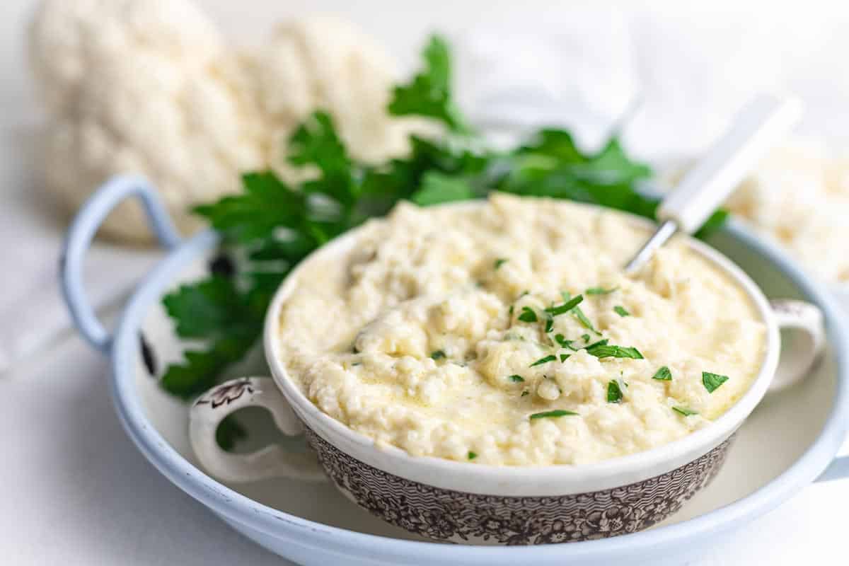 A bowl of creamy mashed cauliflower garnished with chopped parsley, placed on a plate with fresh parsley in the background.