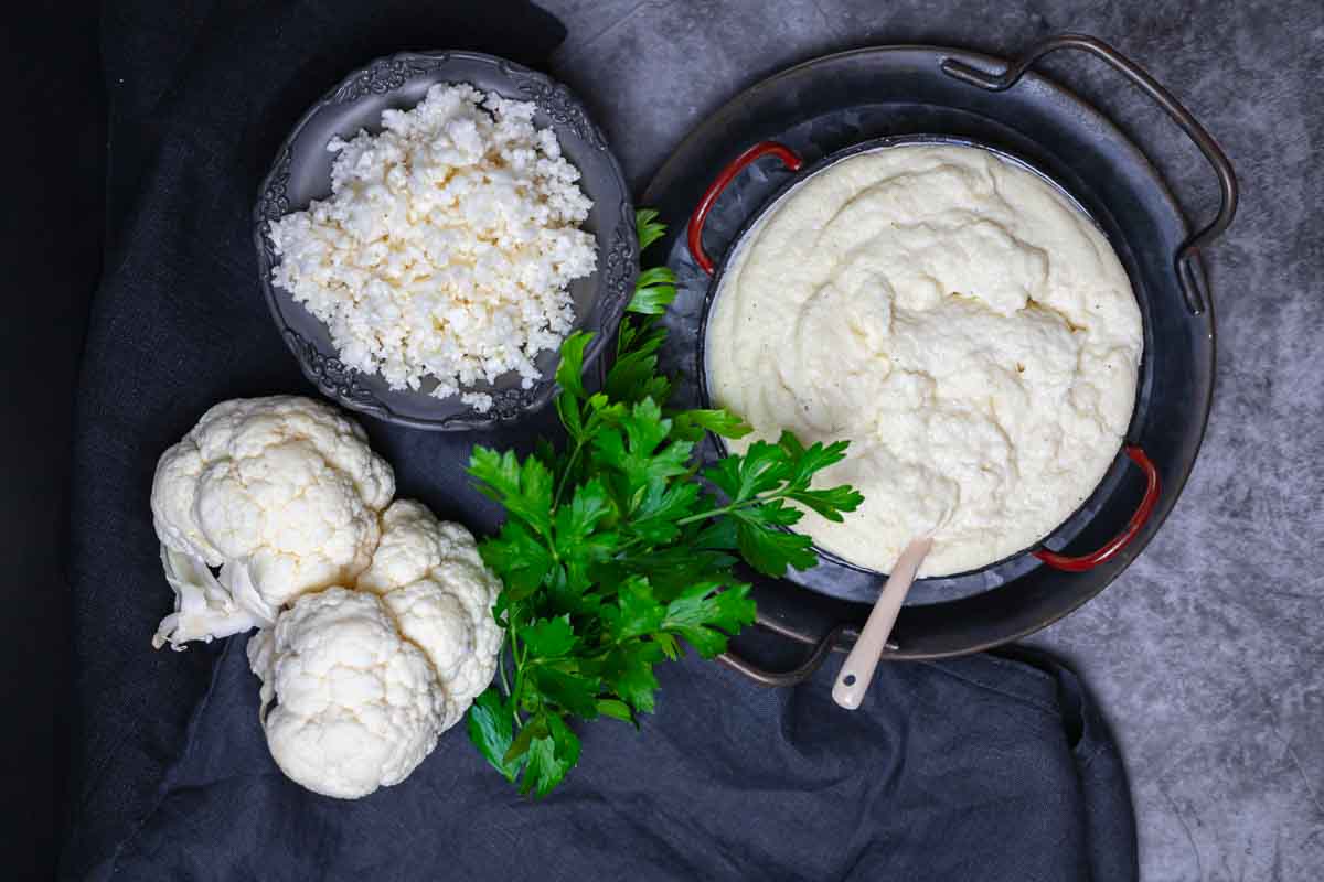 A bowl of cauliflower rice, a dish of mashed cauliflower with a spoon, fresh cauliflower heads, and parsley on a dark surface.
