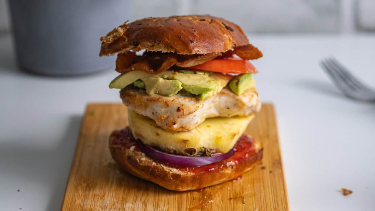 A sandwich with grilled chicken, pineapple, avocado, tomato, bacon, red onion, and sauce on a wooden board, with a pretzel bun.