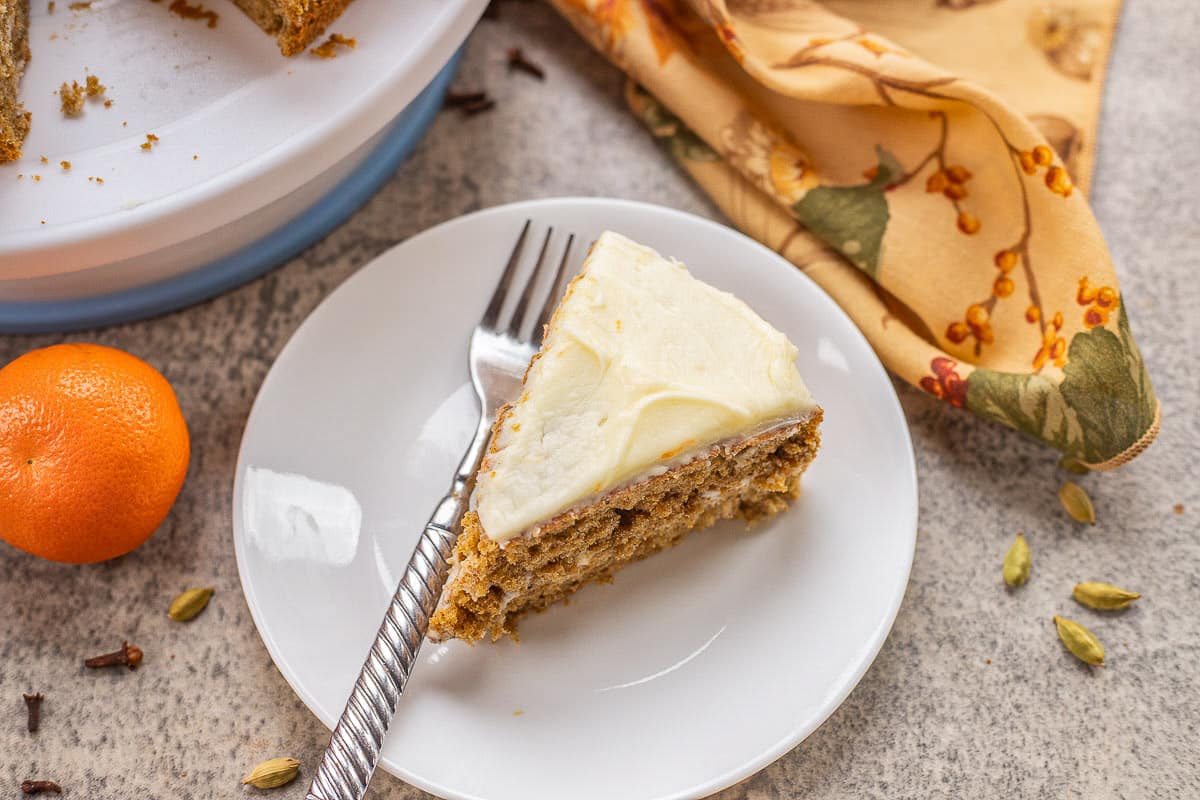 A slice of frosted cake on a white plate with a fork, next to a clementine, a napkin, and scattered cardamom pods and cloves.