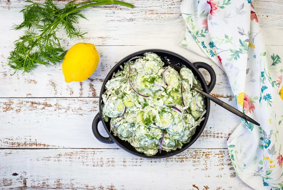 A bowl of creamy cucumber salad with dill and red onion sits on a white wooden table next to a lemon and a floral napkin.