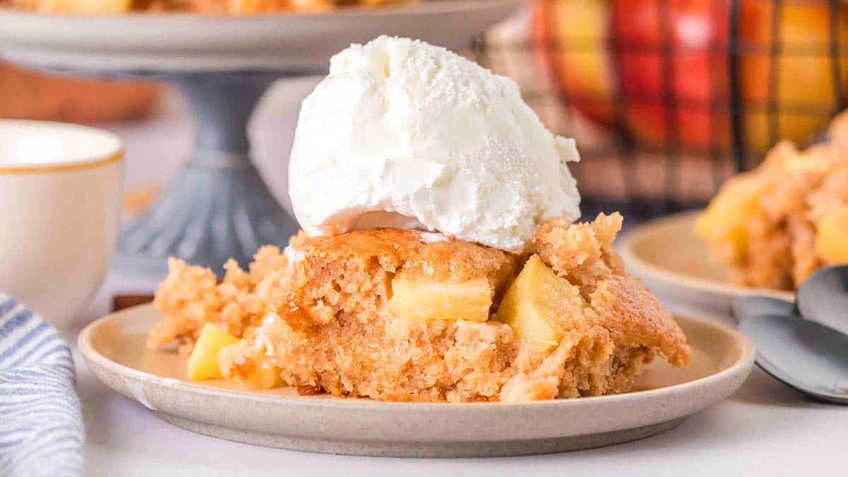 A slice of apple cobbler topped with a scoop of vanilla ice cream is served on a plate, with more cobbler and apples visible in the background.