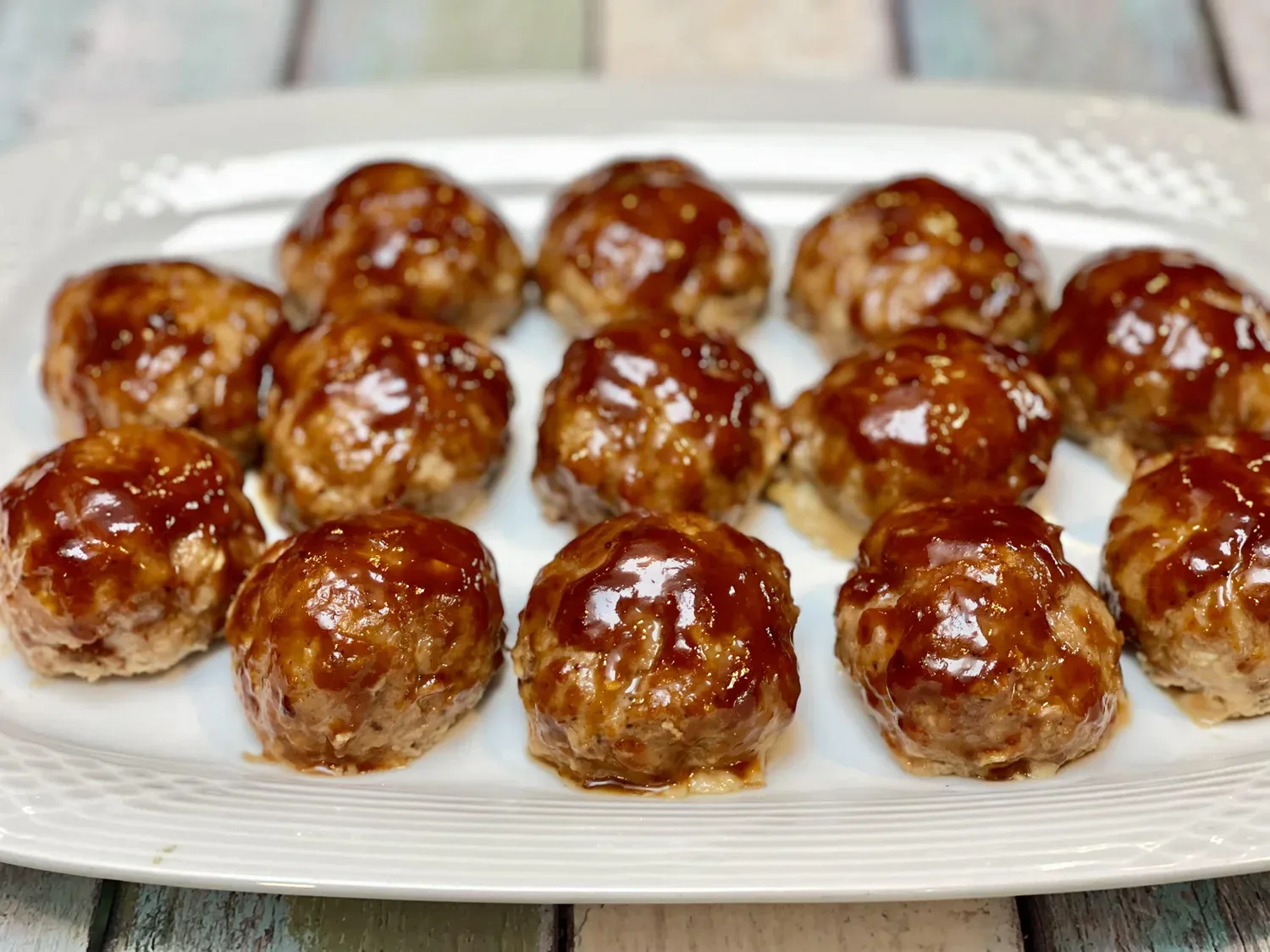 Fourteen glazed meatballs are arranged neatly on a white rectangular plate, displayed on a light wooden surface.