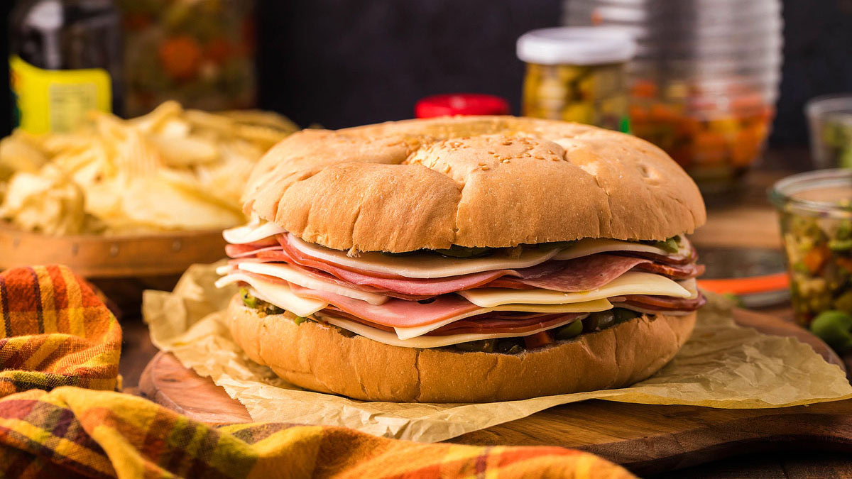 Large sandwich with multiple layers of deli meats and cheeses on a round sesame bun, placed on parchment paper with chips and jars of pickles in the background.