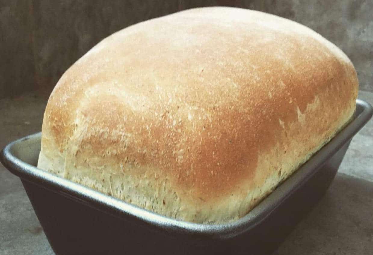A loaf of freshly baked bread sits in a rectangular metal baking pan on a gray surface.