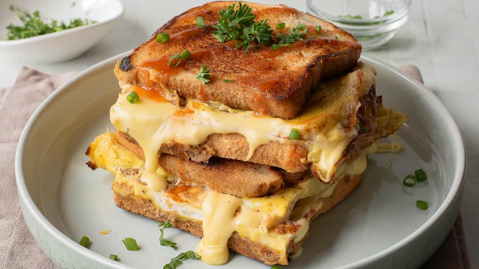 Two stacked grilled cheese and egg sandwiches on a plate, with melted cheese, chopped parsley, and green onions on top.