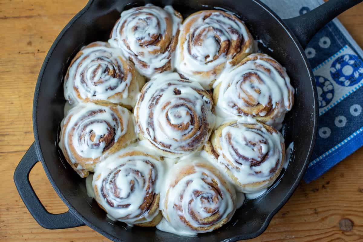 A cast iron skillet filled with nine frosted cinnamon rolls, sitting on a wooden surface next to a blue and white patterned cloth.