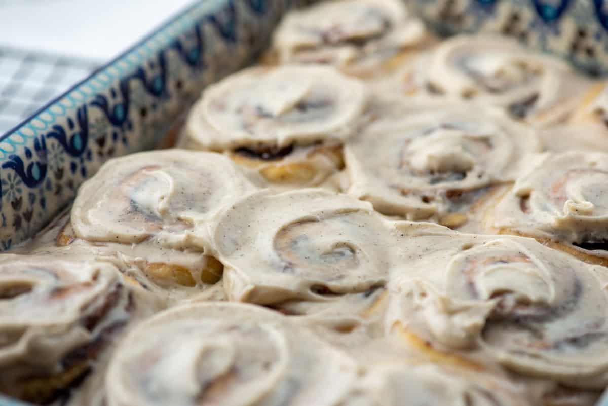 A close-up of cinnamon rolls in a patterned baking dish, topped with a thick layer of cream cheese frosting.