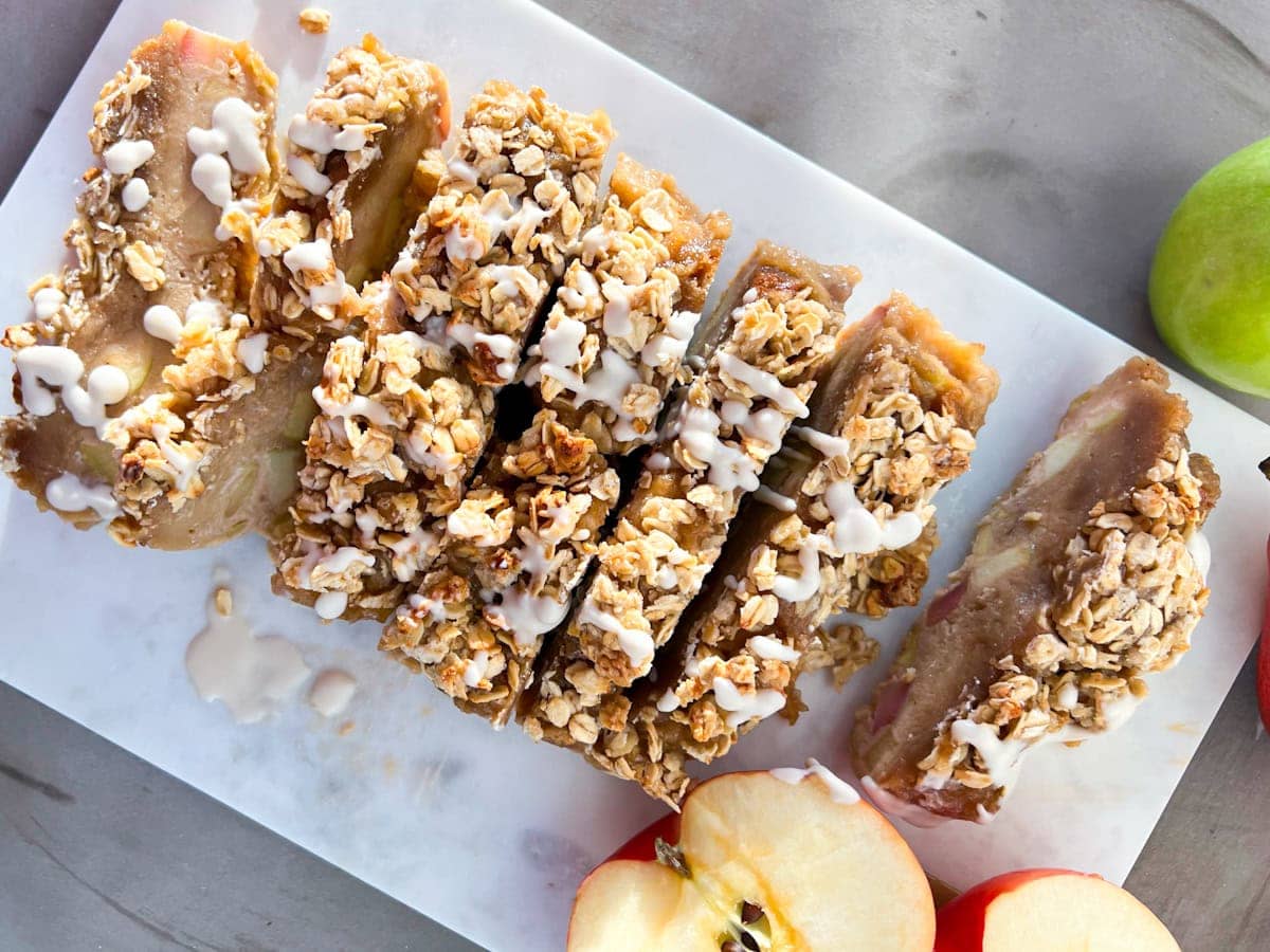 Sliced apple oat bars with icing drizzled on top, displayed on a white cutting board next to fresh apples.