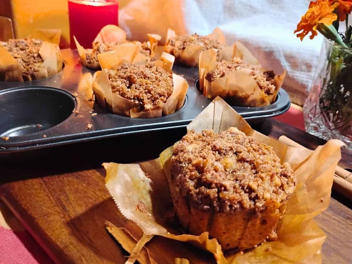A tray of freshly baked muffins with crumb topping, one placed on a wooden board, next to a lit candle and a small vase with flowers.