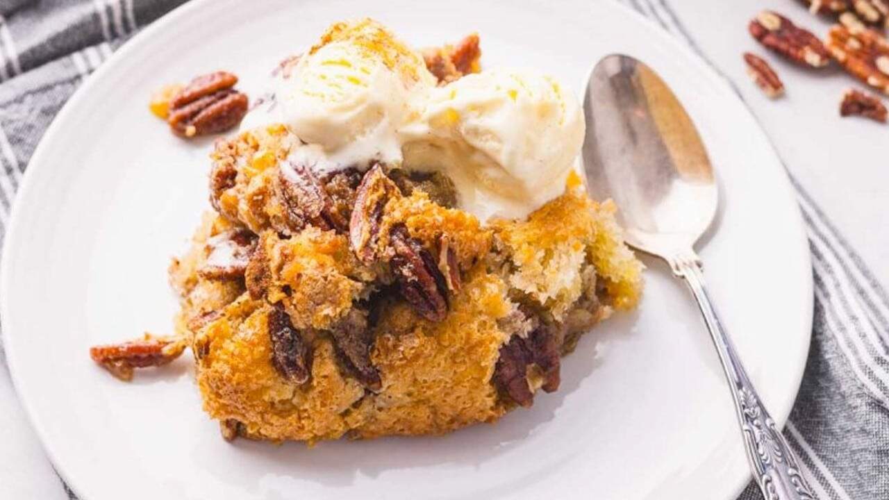 A serving of pecan cobbler topped with vanilla ice cream on a white plate with a spoon next to it.