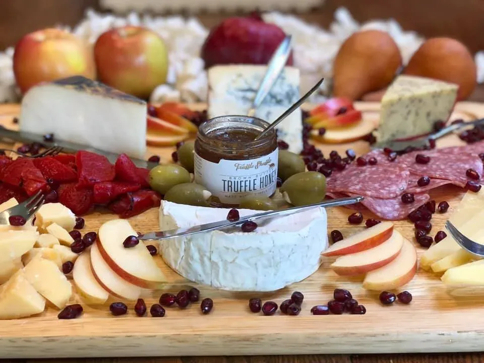 A wooden board with assorted cheeses, sliced apples, pears, strawberries, olives, salami, pomegranate seeds, and a jar of truffle honey in the center.