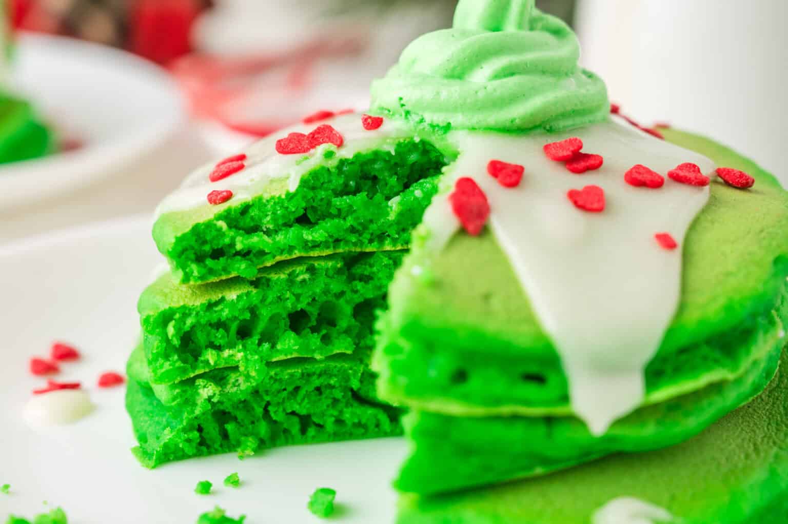A stack of green pancakes with white frosting, red heart-shaped sprinkles, and a frosted swirl on top, partially cut to show the inside.