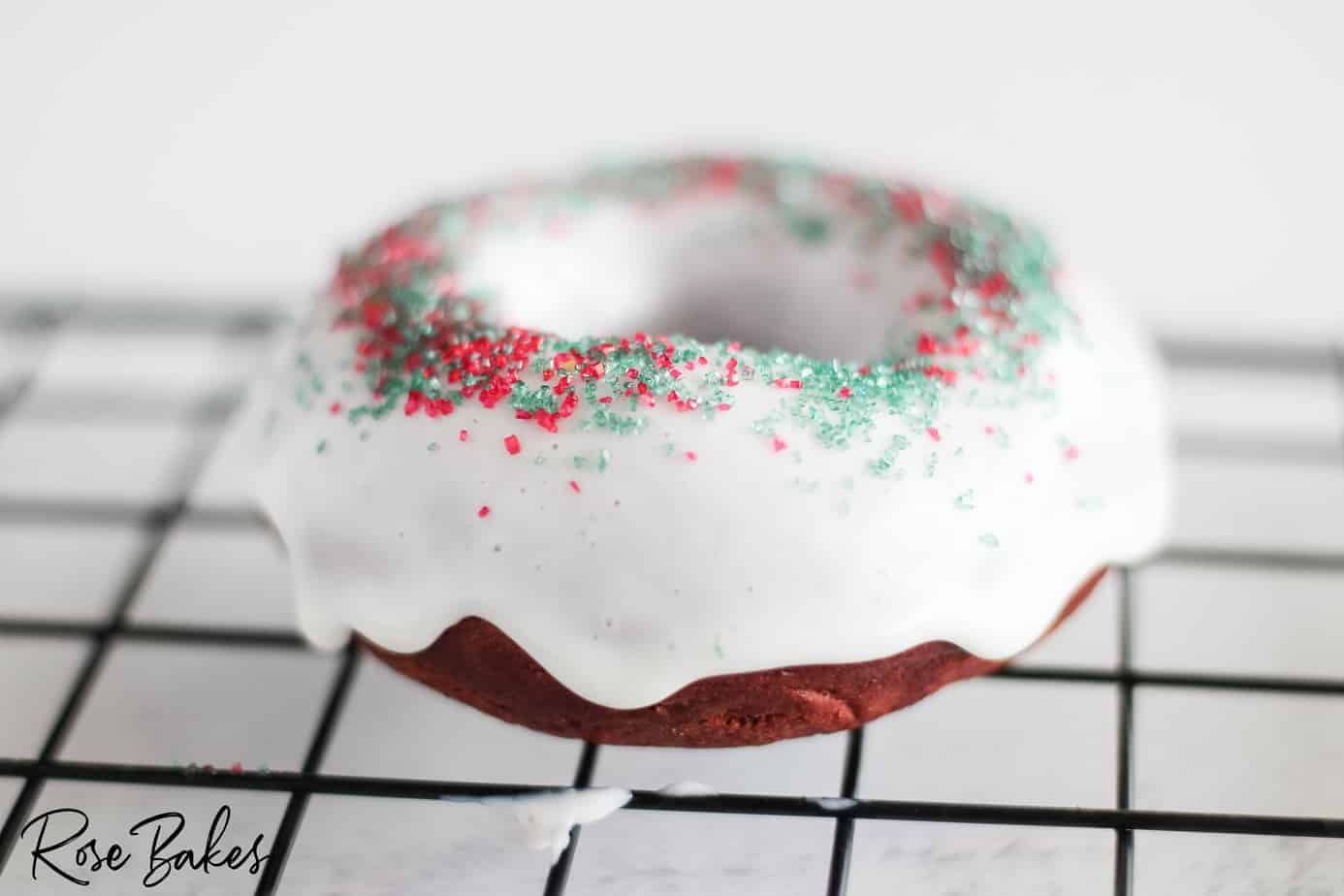 A chocolate donut with white icing and red and green sprinkles rests on a black cooling rack.