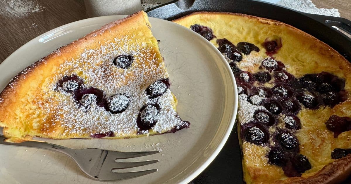 A slice of blueberry Dutch baby pancake dusted with powdered sugar on a plate with a fork, next to the remaining pancake in a cast iron skillet.