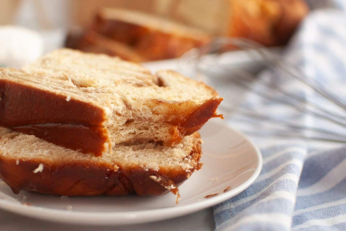 Two slices of cinnamon swirl bread are stacked on a white plate, with a whisk and striped cloth in the background.