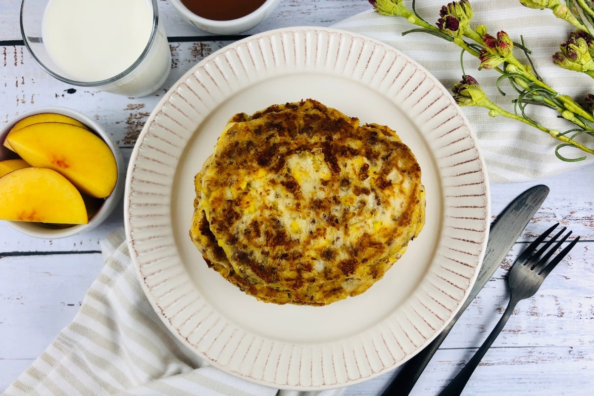 A stack of pancakes on a plate with a fork and knife, next to a glass of milk, peach slices, and flowers on a white wooden table.